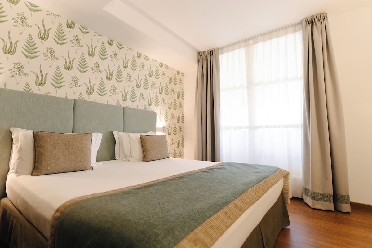 Ein ordentlich bezogenes Bett mit einer grün-beigen Tagesdecke, ein Kopfteil mit Tapete mit Blattmuster und ein Fenster mit beigen Vorhängen.
