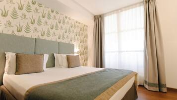 Ein ordentlich bezogenes Bett mit einer grün-beigen Tagesdecke, ein Kopfteil mit Tapete mit Blattmuster und ein Fenster mit beigen Vorhängen.