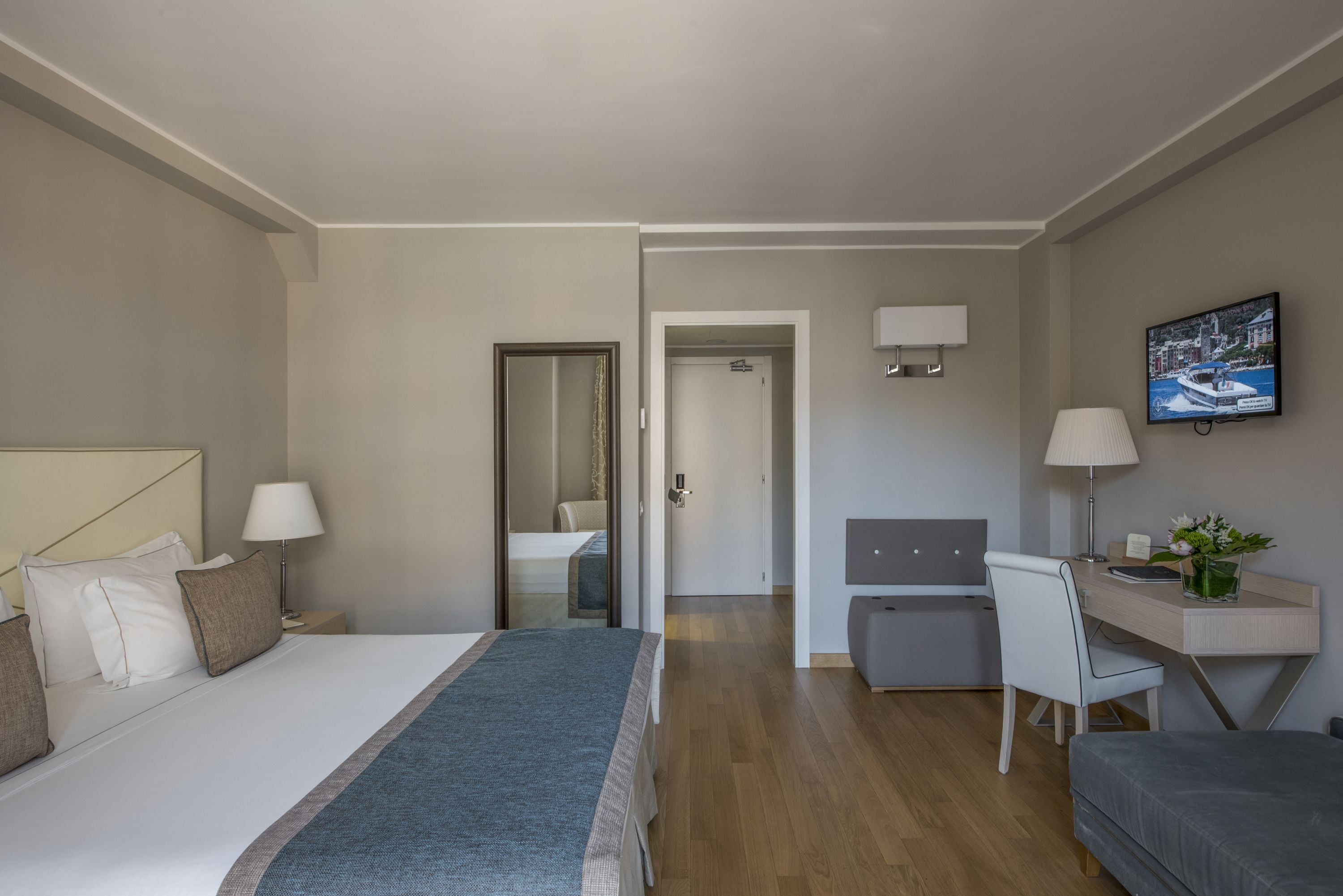 Una habitación de hotel con cama, mesita de noche, lámpara, escritorio, silla y televisor en la pared.