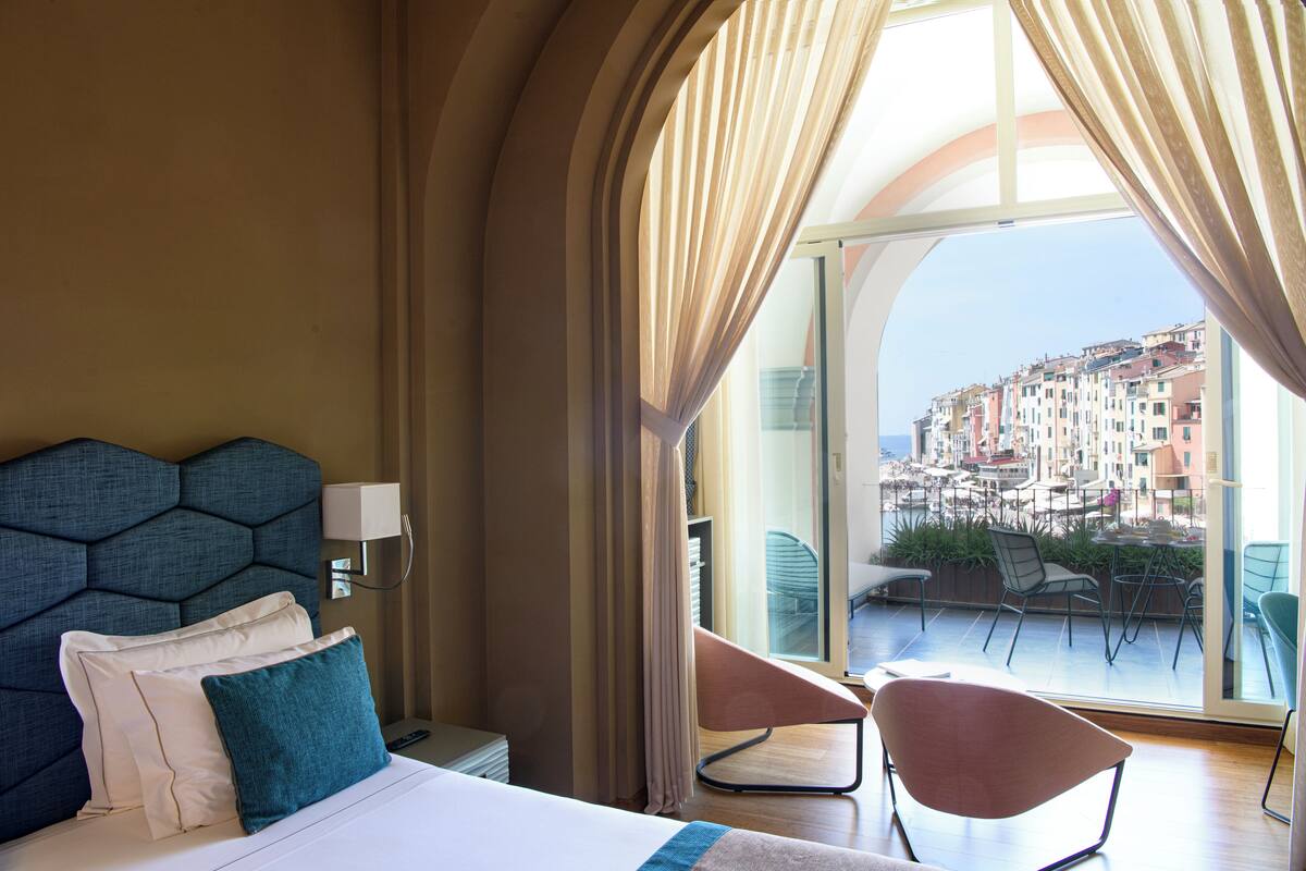 Ein Hotelzimmer mit einem großen Bett, Blick auf eine Küstenstadt und einem Balkon mit Sitzgelegenheiten.