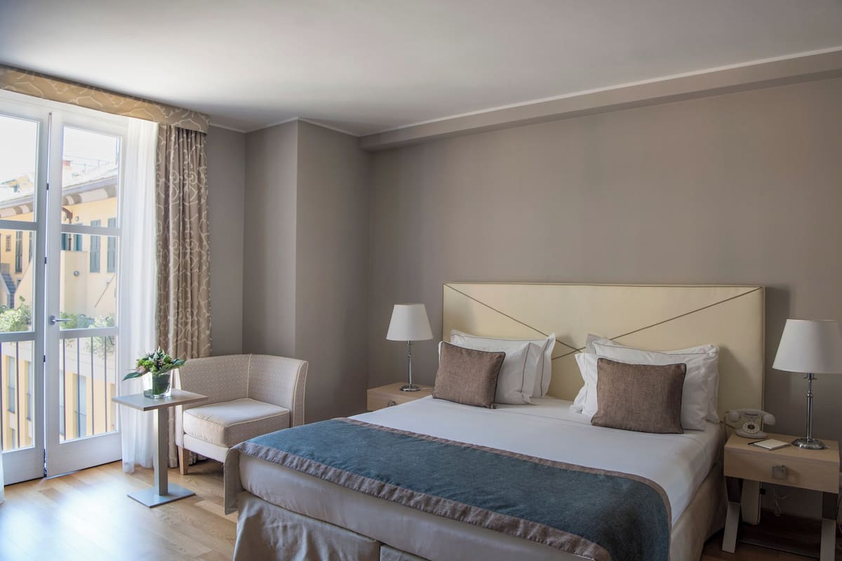 Ein Hotelzimmer mit einem großen Bett, zwei Nachttischen, einem kleinen Tisch mit einer Lampe, einem Sessel und Blick auf Gebäude durch ein großes Fenster.