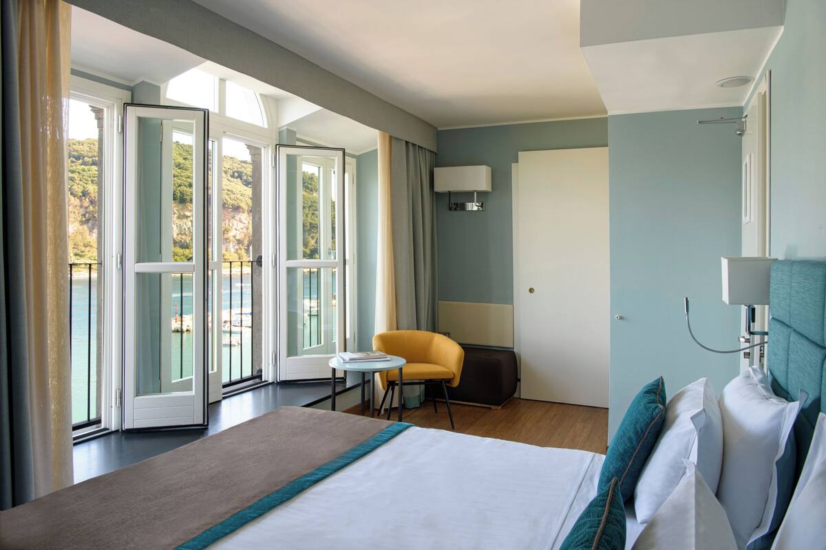 Ein Hotelzimmer mit einem großen Bett, einem kleinen Tisch, einem Sessel und Blick ins Freie durch große Fenster.