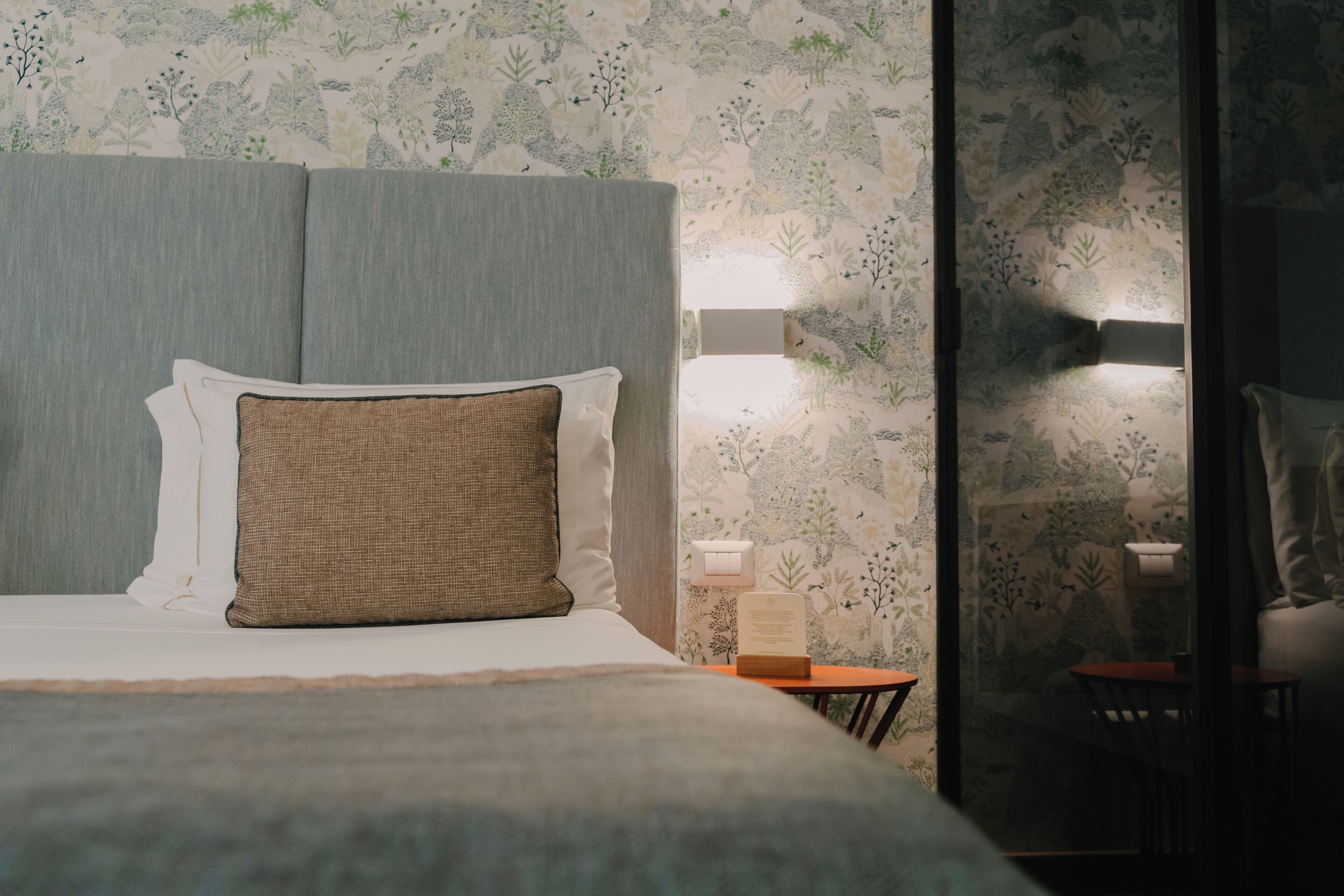 Un lit bien fait, avec une tête de lit, une table de chevet avec une lampe, et une petite table entourée de chaises au fond de la pièce.