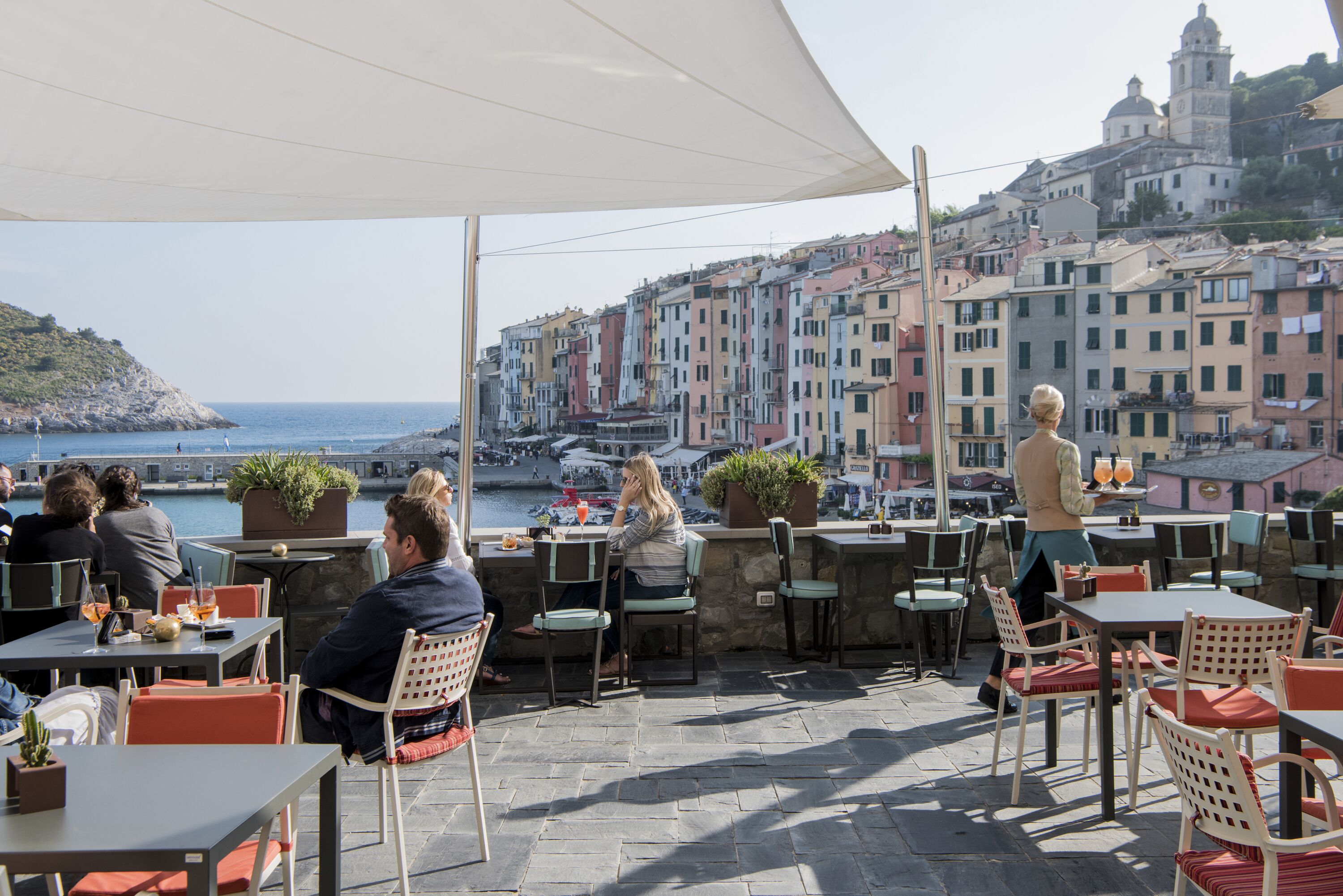 Un caffè all'aperto con tavoli e sedie, con vista su una cittadina costiera e palazzi colorati.