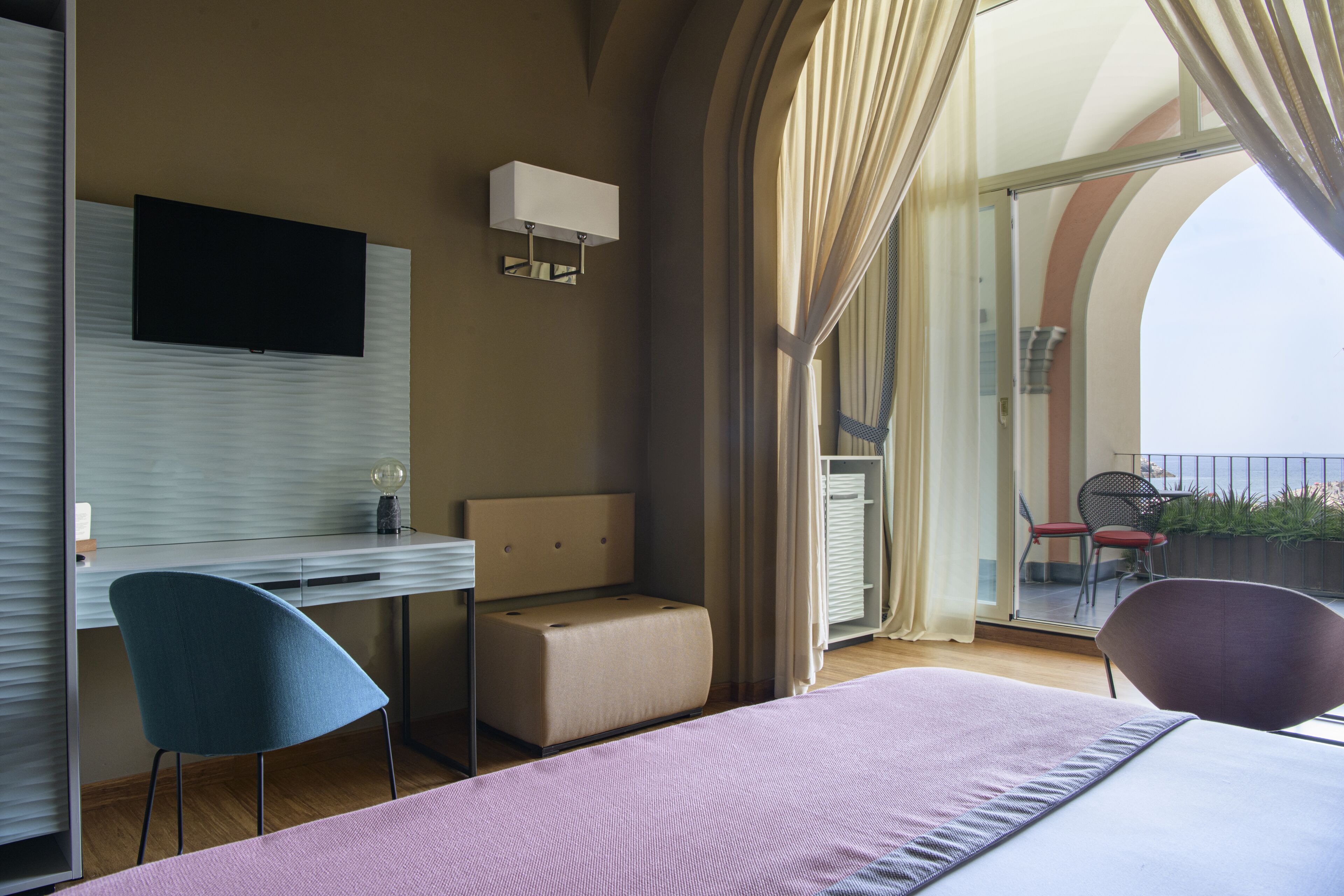 Quarto de hotel moderno com cama, escrivaninha, cadeira, televisão e varanda com cadeira e plantas.