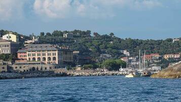 A coastal town with a prominent hotel featuring a large central building with multiple windows and balconies, situated near a rocky shoreline with boats and a clear view of the sea.