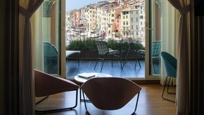 Habitación de hotel con un amplio ventanal de arco, un balcón con sillas y vistas a un colorido paisaje urbano.