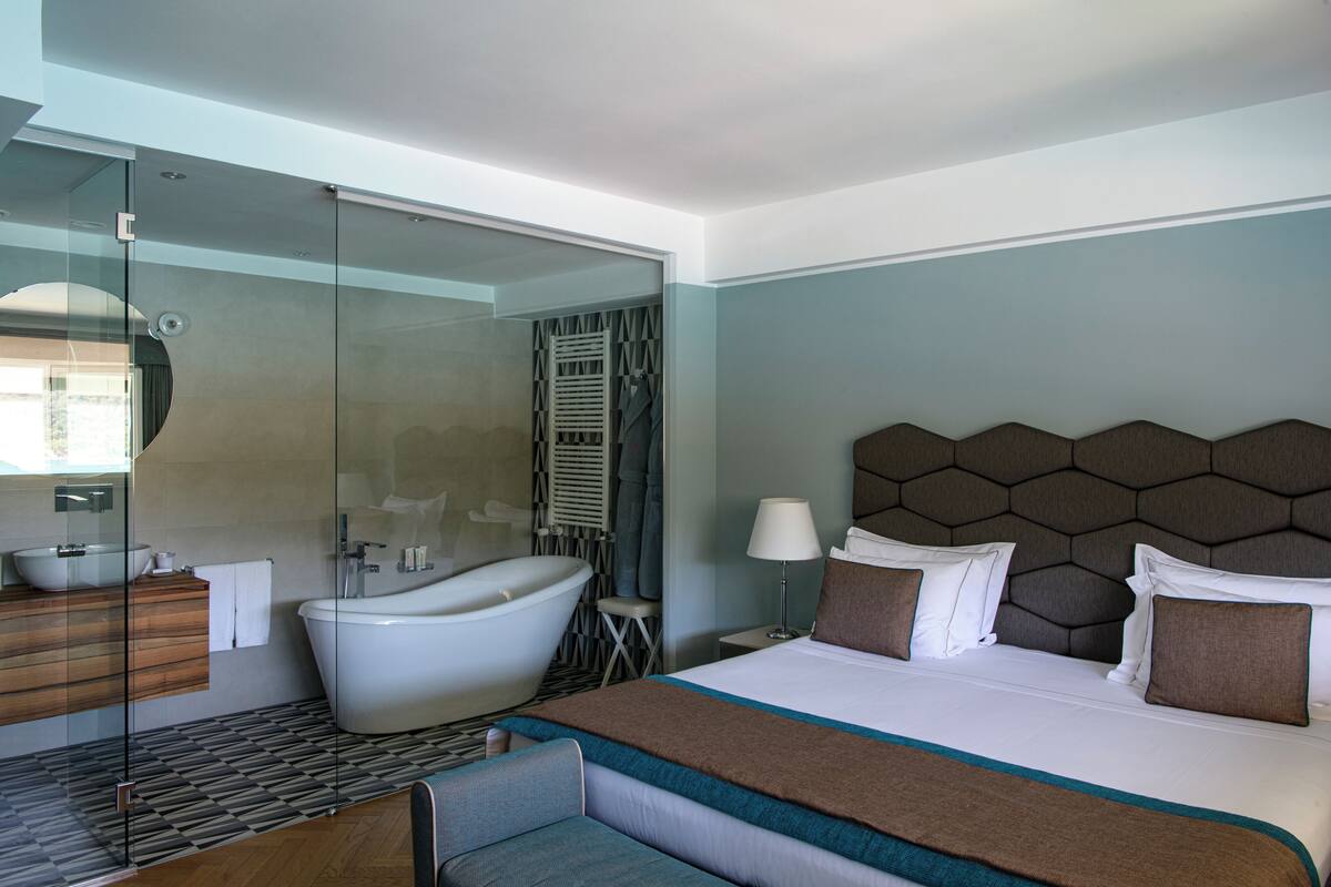 Ein modernes Hotelzimmer mit einem großen Bett, einer gläsernen Dusche, einer Badewanne, einem Holznächtigungstisch und einem Nachttisch mit Lampe.