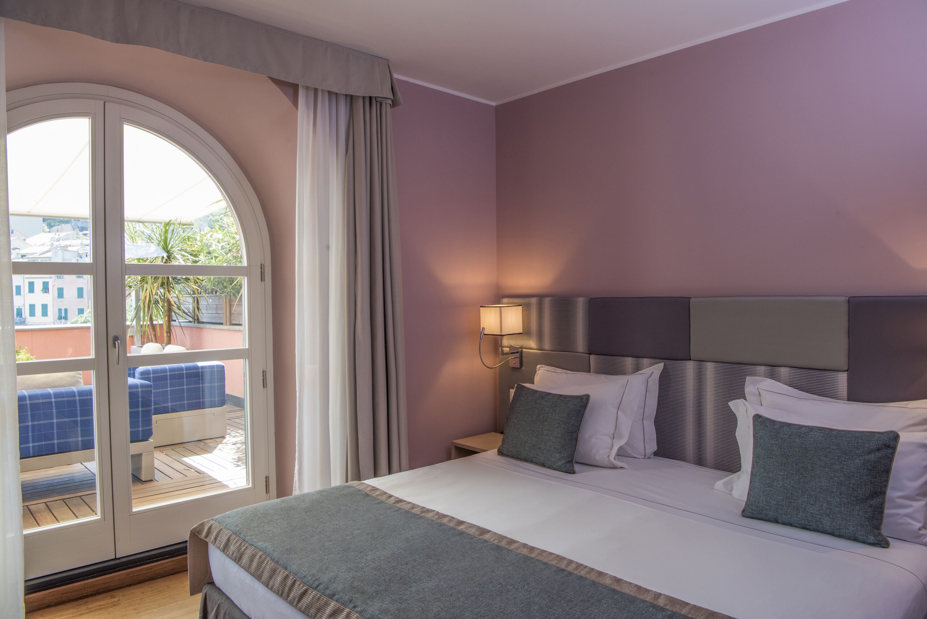A hotel room with a bed, a nightstand, a lamp, a view through a glass door to an outdoor seating area, and a balcony.