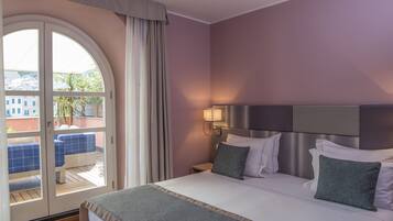 Ein Hotelzimmer mit Bett, Nachttisch, Lampe und Blick durch eine Glastür auf einen Sitzbereich im Freien sowie einen Balkon.