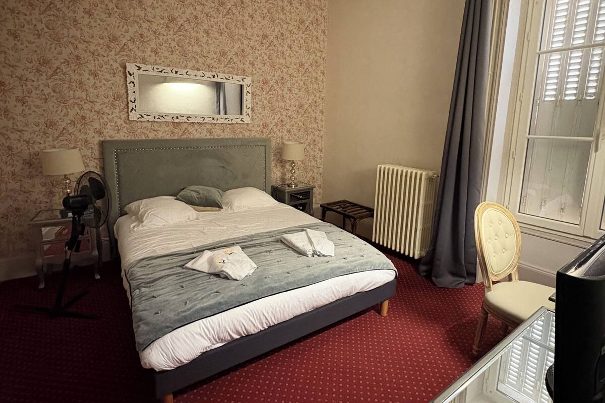 Habitación de hotel con cama, silla, televisor, espejo, ventana con cortinas y radiador.
