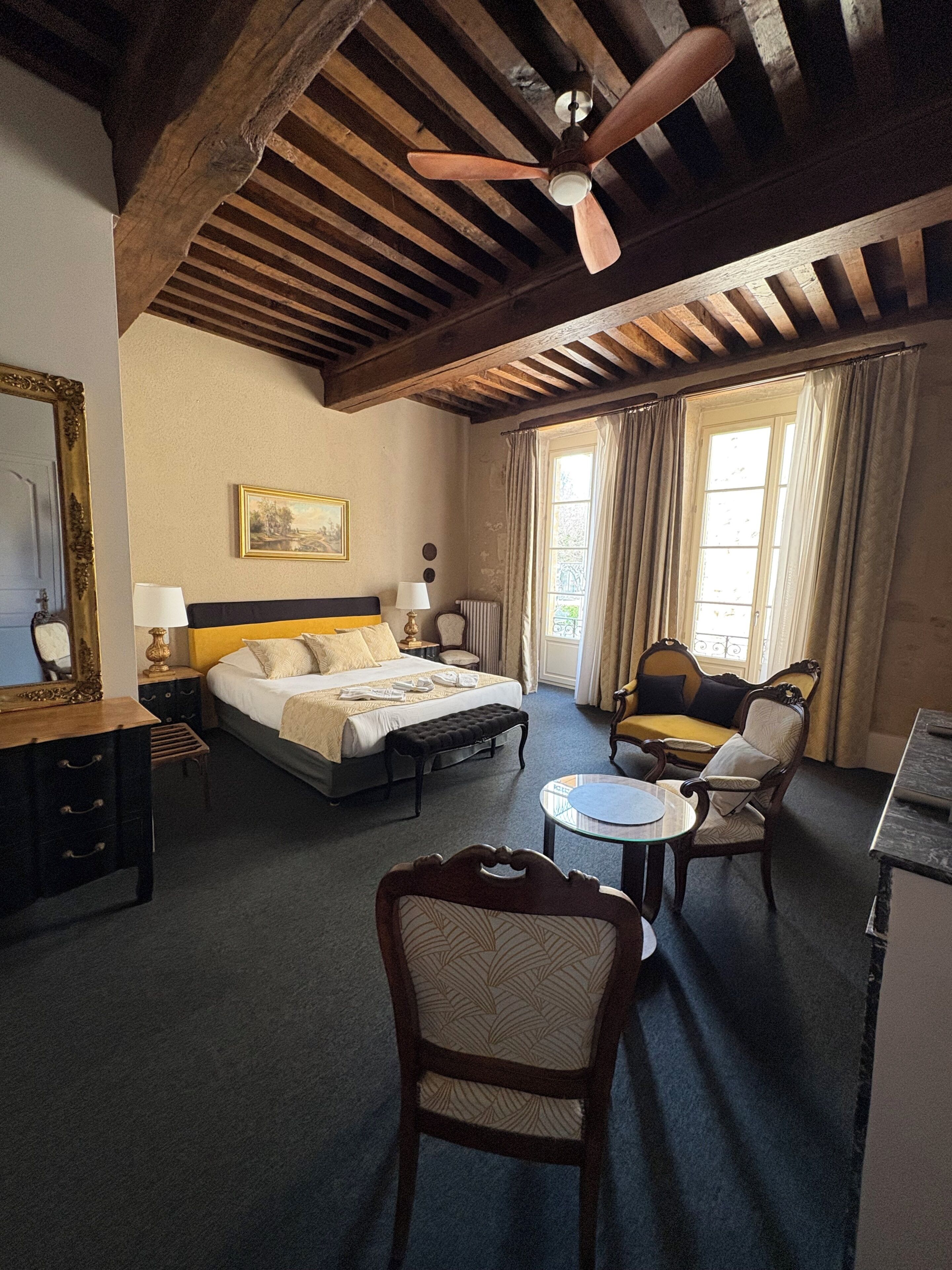 A hotel room with a bed, a chair, a small table, a ceiling fan, a window with curtains, and a radiator.