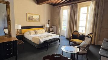 A hotel room with a bed, a chair, a small table, a ceiling fan, a window with curtains, and a radiator.