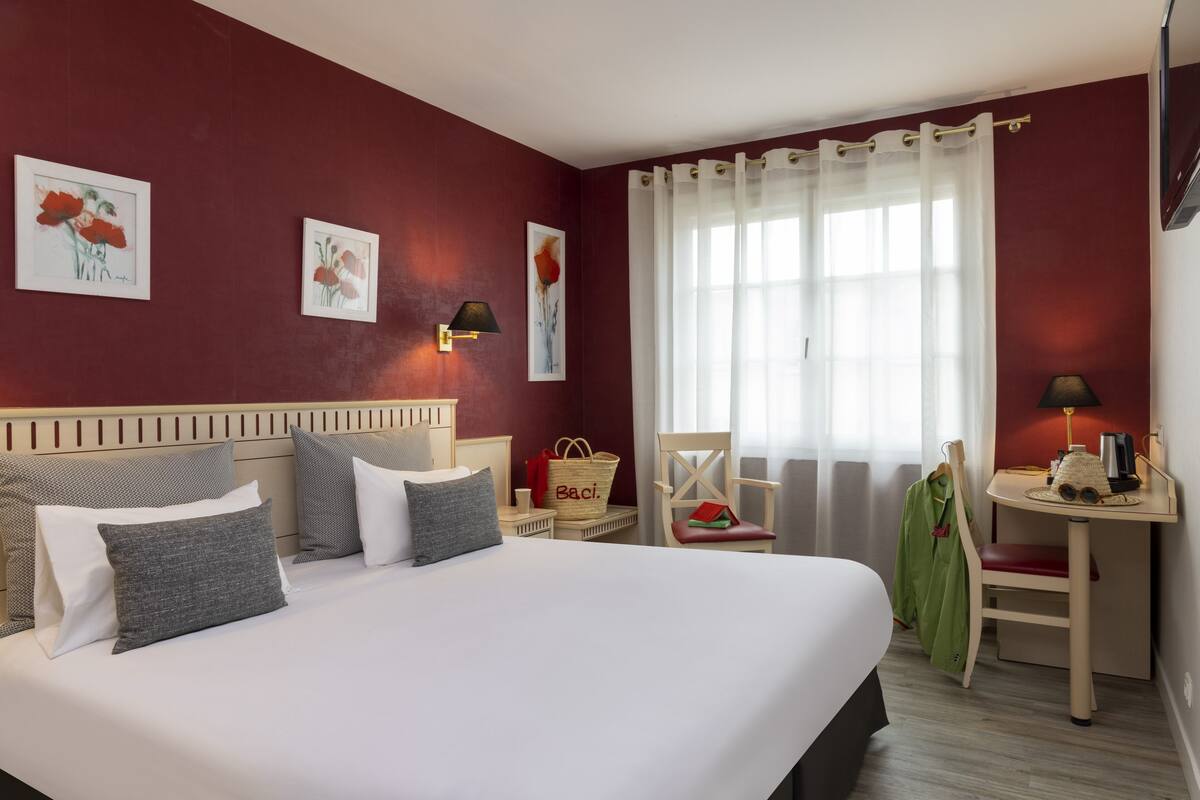 Quarto de hotel com uma cama grande, uma escrivaninha com luminária, uma cadeira, uma mesinha e obras de arte nas paredes.