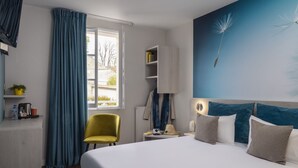 Habitación de hotel con cama, una silla amarilla, un escritorio con lámpara, repisas con artículos de aseo y una ventana con cortinas.