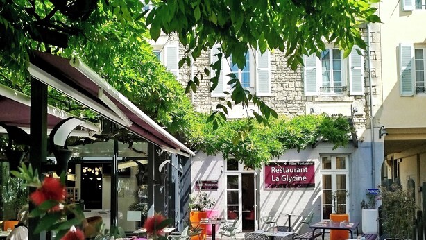 Un café al aire libre con mesas y sillas, rodeado de vegetación y edificios.