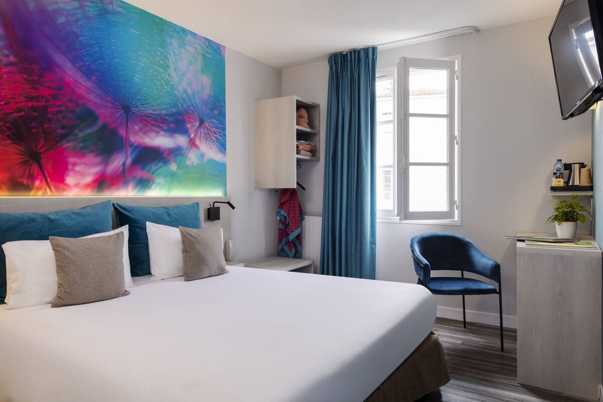 Quarto de hotel com uma cama grande, uma escrivaninha com cadeira, uma televisão fixada na parede e uma janela com cortinas.