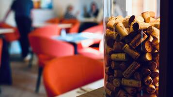 A glass jar filled with wine corks on a wooden table in a modern, brightly lit room with orange chairs and a blurred background of people.