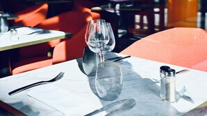 Un ambiente de restaurante con una mesa lista para dos personas, que incluye una copa de vino, cubiertos y saleros y pimenteros.