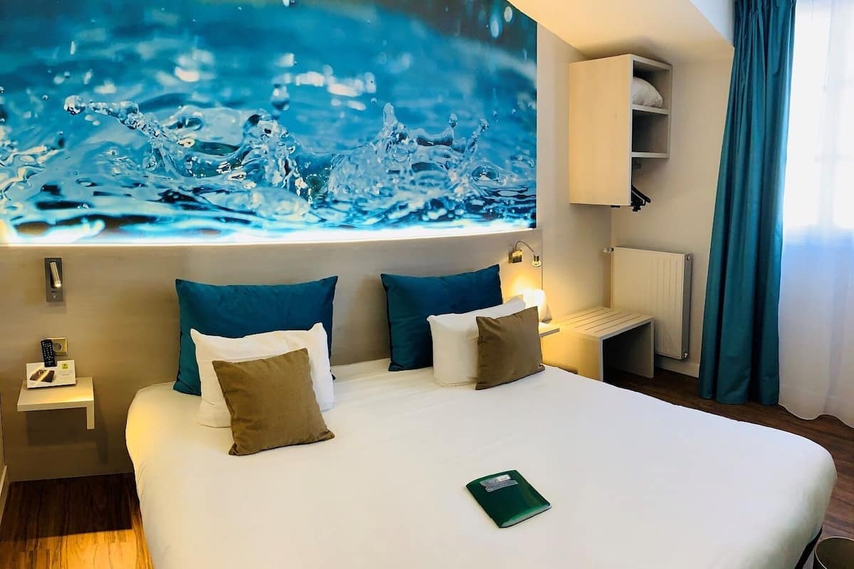 A hotel room with a large bed, two nightstands, a bedside table, a wall-mounted TV, a radiator, and a window with curtains.