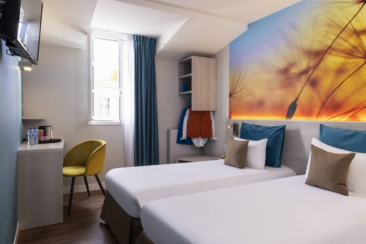 Quarto de hotel com duas camas, uma escrivaninha, uma cadeira, um guarda-roupa e uma janela grande com cortinas.