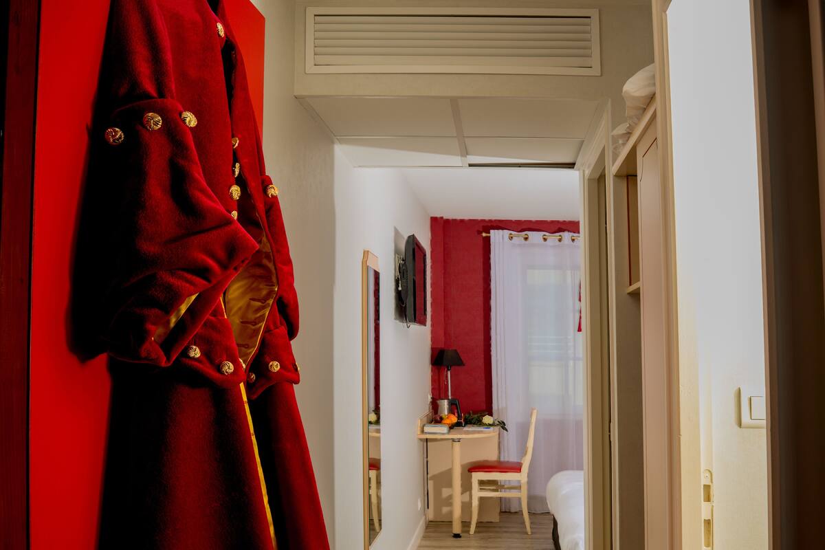 Quarto de hotel com roupão vermelho pendurado na parede, uma escrivaninha com abajur, uma cadeira e uma cama.