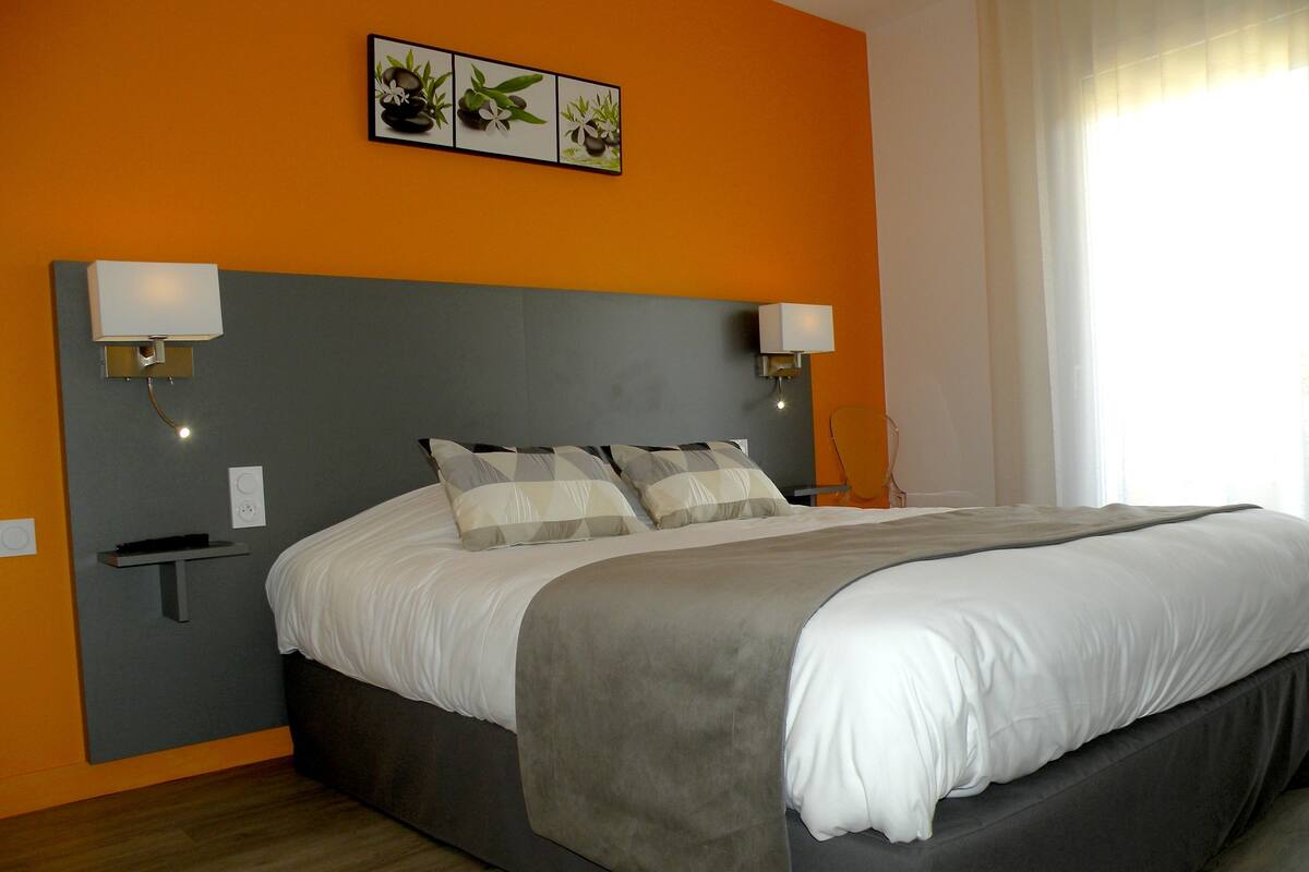 A modern bedroom with a large bed, grey headboard, and orange walls. There are two bedside lamps and a small shelf.