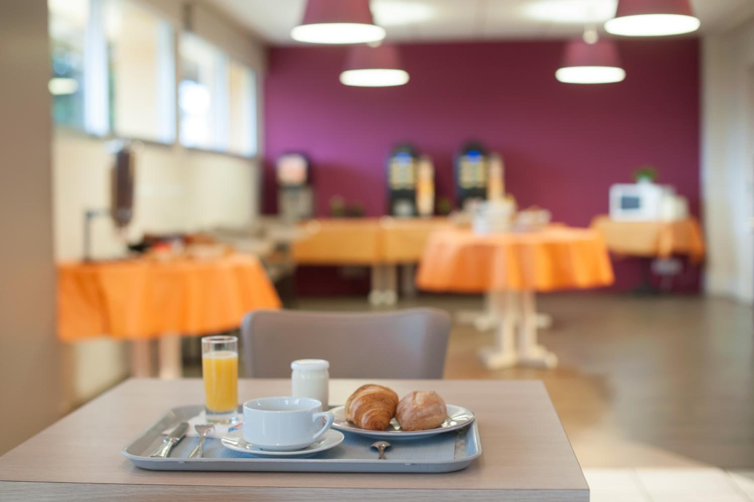 Un plateau de petit-déjeuner avec des croissants, un verre de jus d’orange et une tasse de café, posé sur une table dans un coin repas aux tables orange et aux murs violets.