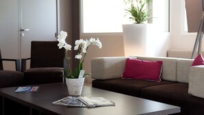 Een moderne woonkamer met een bank, een bijzettafeltje met tijdschriften, een potorkidee en een groot raam met uitzicht op beplanting.