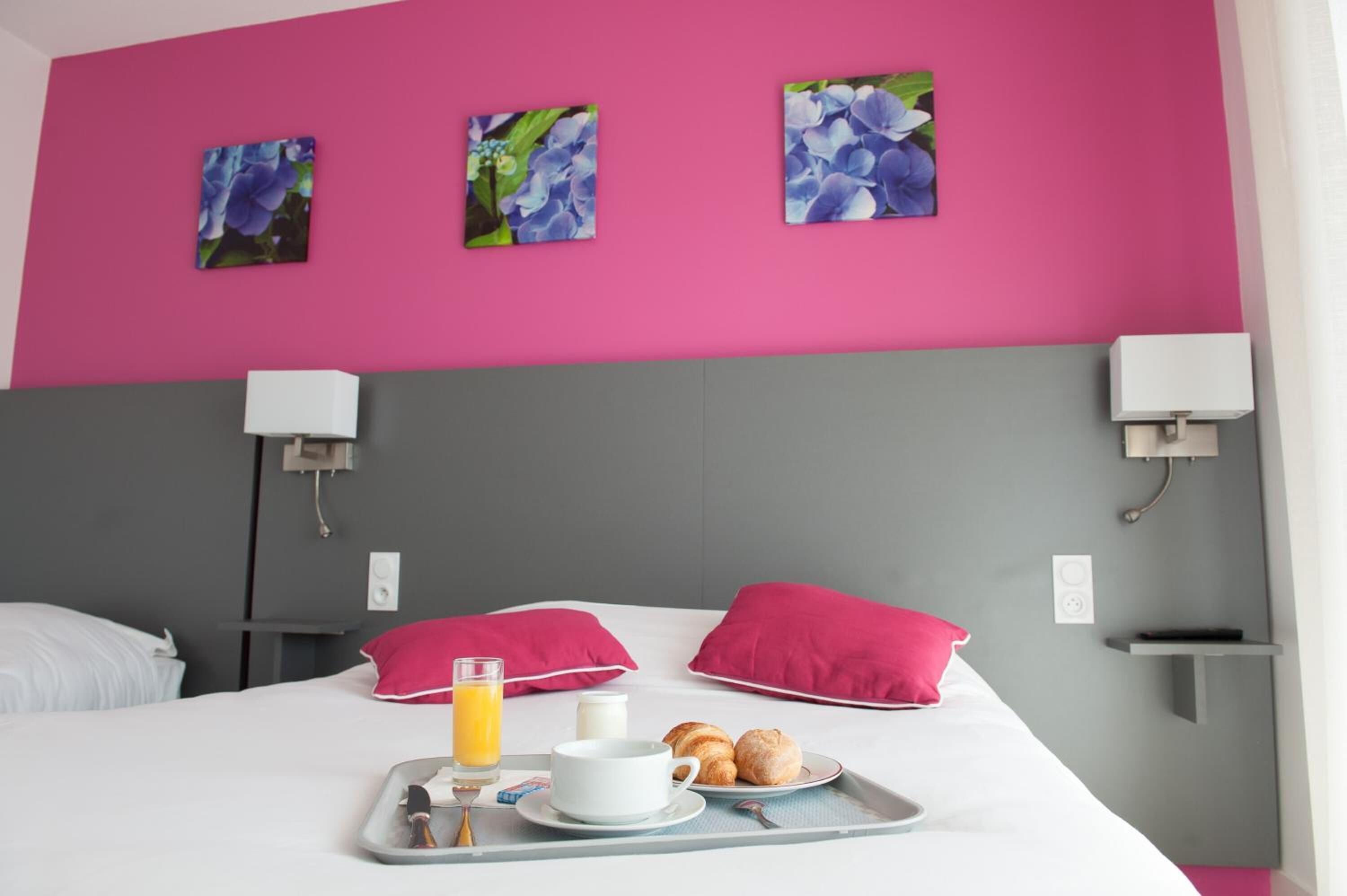 Une chambre d’hôtel avec un lit, des oreillers roses, un plateau avec des croissants, une tasse et un verre de jus d’orange.