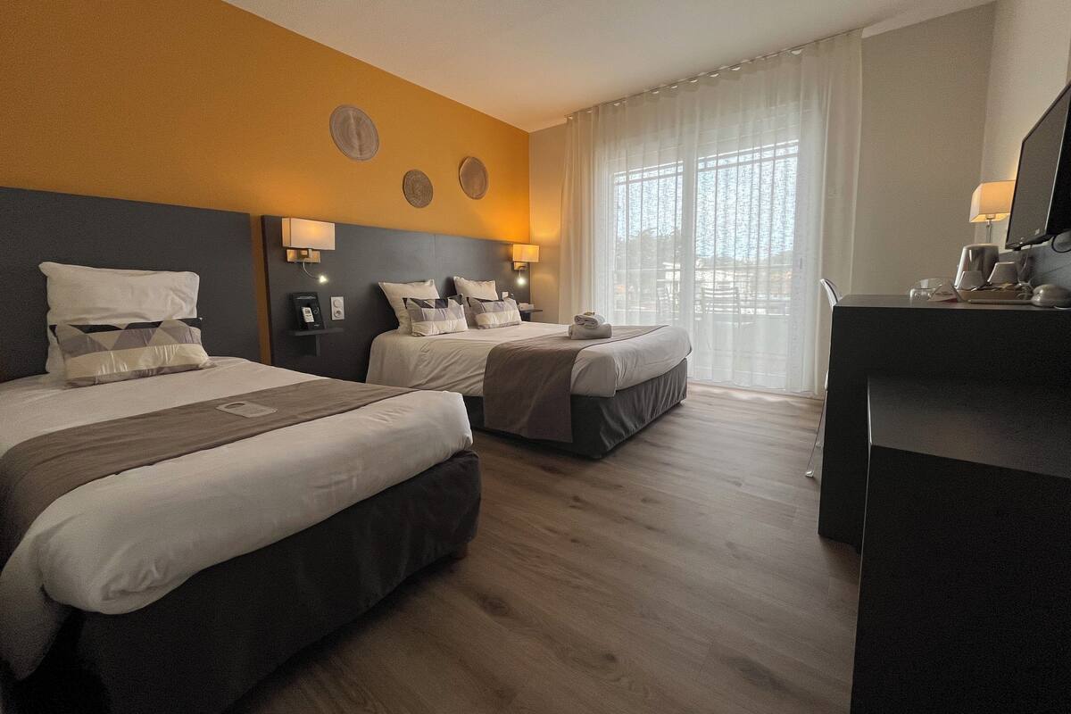 A hotel room with two beds, a desk, and a large window with sheer curtains.