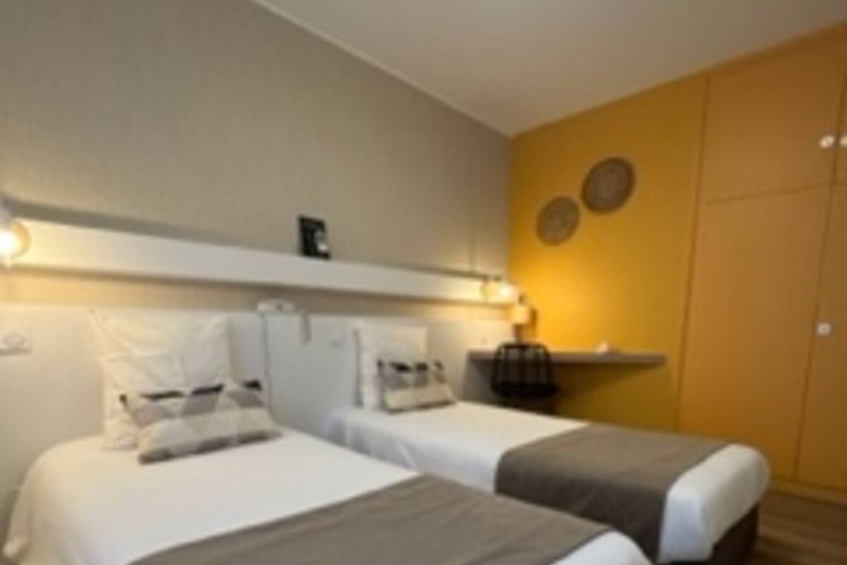 A hotel room with two beds, a small table, a wall-mounted shelf, and a ceiling light fixture.