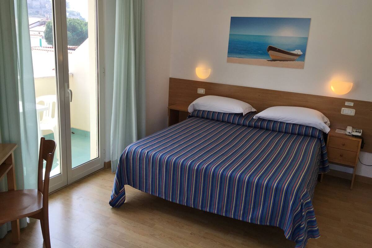 Ein Hotelzimmer mit Bett, Nachttisch, Stuhl, einem an der Wand hängenden Gemälde eines Bootes und Blick durch das Fenster.