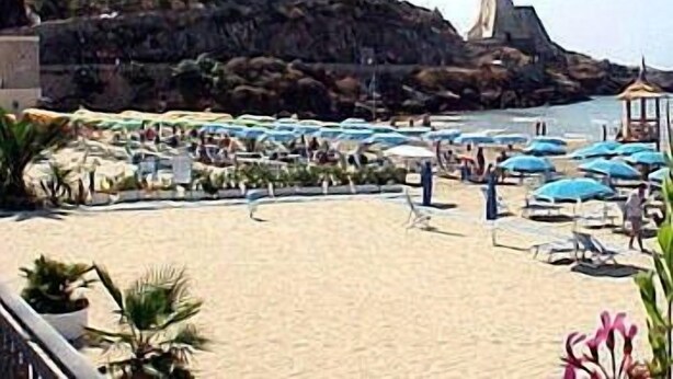 Uma praia com guarda-sóis e espreguiçadeiras, um penhasco rochoso e edifícios ao fundo.
