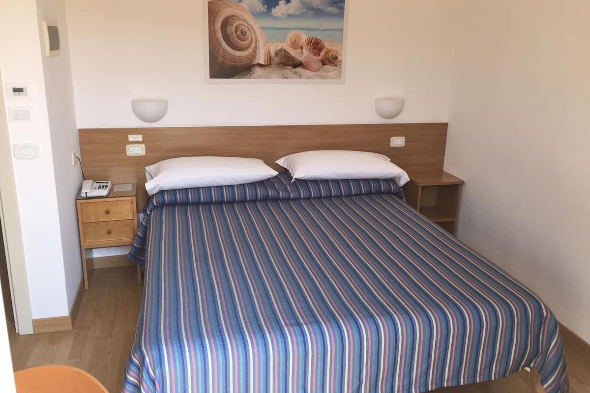 Ein Hotelzimmer mit einem Bett, zwei Nachttischen, einem Schreibtisch, einem an der Wand montierten Telefon und einem Bild von Meeresschalen.