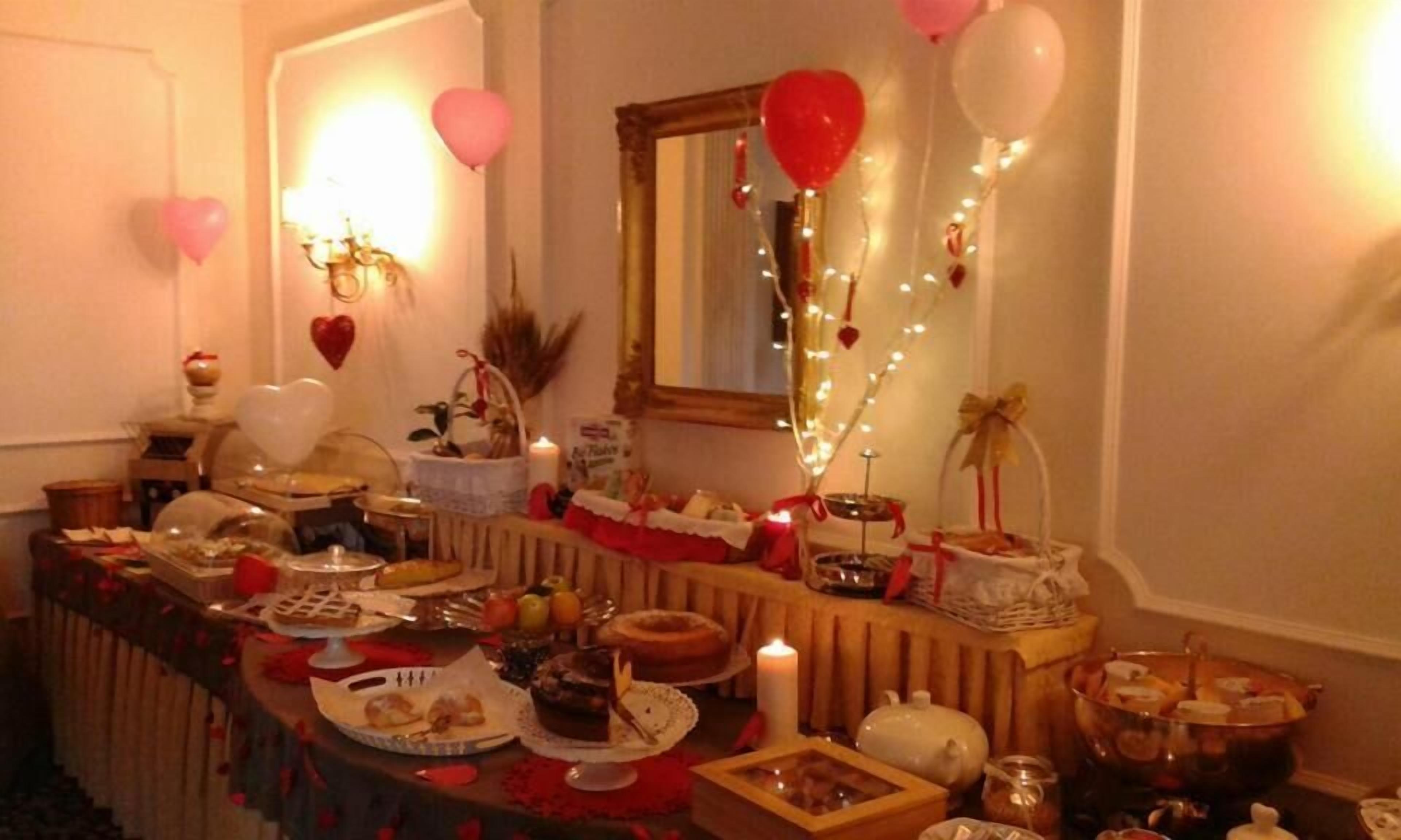 Un comedor con una larga mesa dispuesta para una comida, decorado con globos con forma de corazón y luces colgantes.