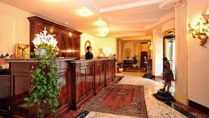 A lobby with a reception desk, a large mirror, a statue, and a patterned carpet. - Hotel Royal (Bolsena)