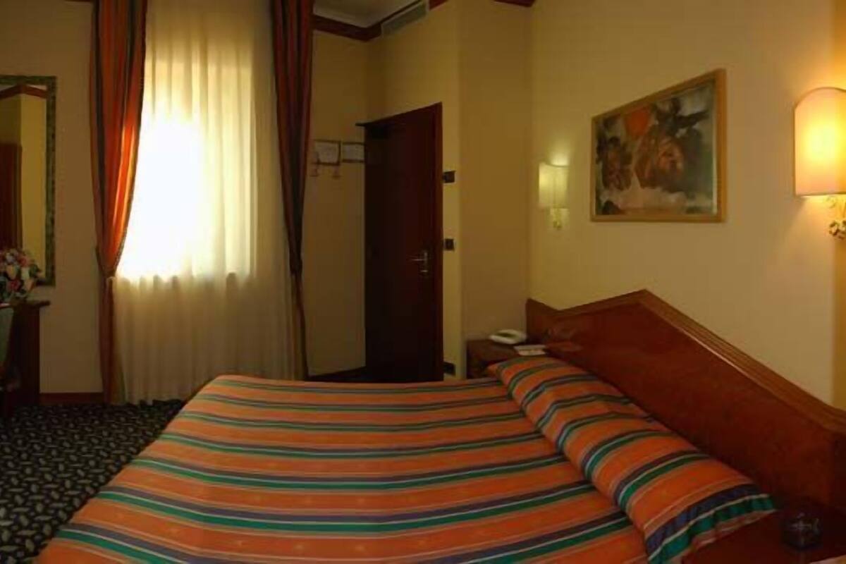 A hotel room with a bed, curtains, a painting, and a lamp.