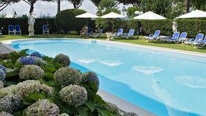 A swimming pool surrounded by blue chairs and umbrellas, with trees and hydrangeas in the foreground. - Hotel Royal (Bolsena)