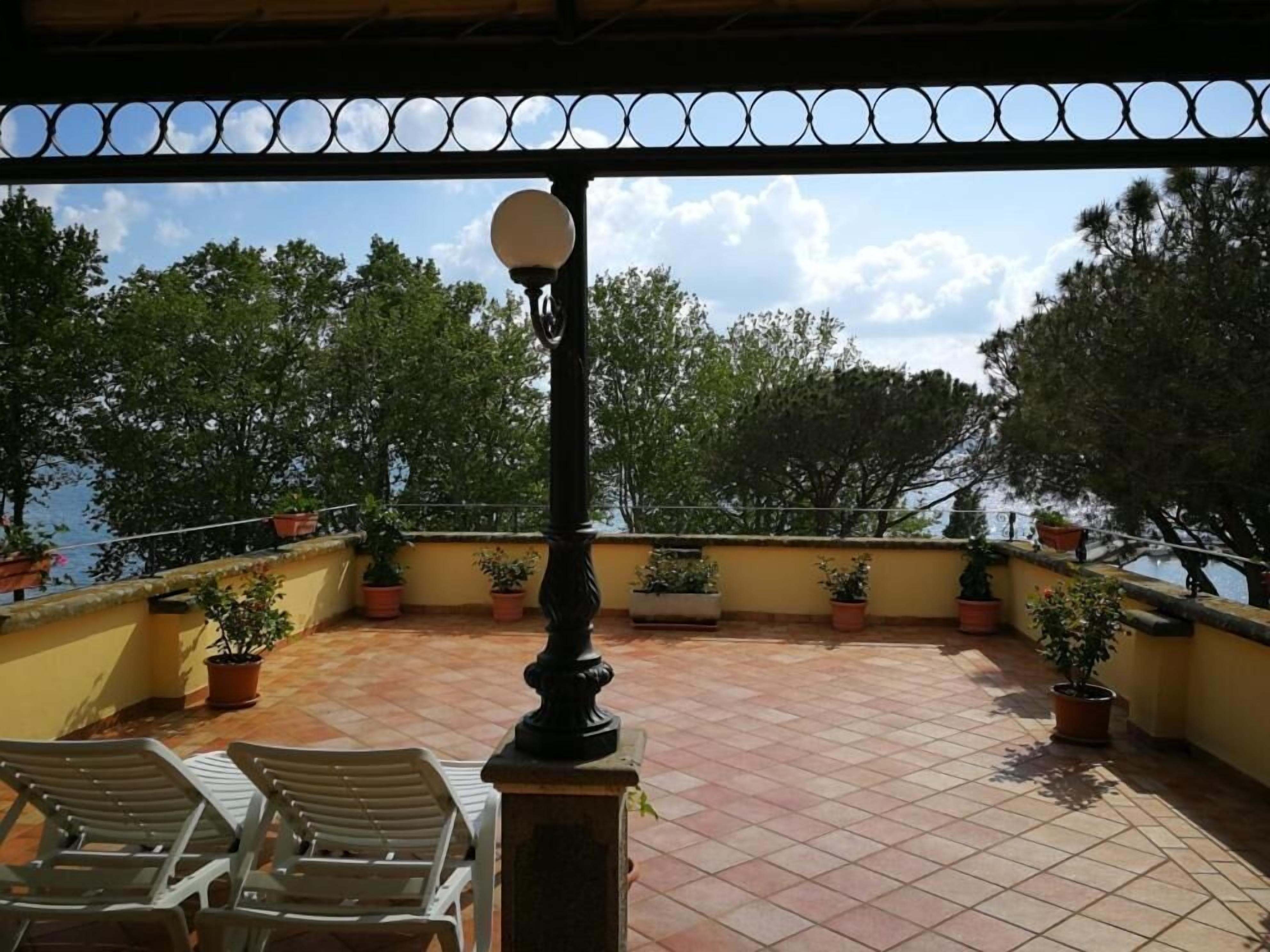 Une terrasse agrémentée de plantes en pot, d’un lampadaire et de deux chaises en plastique blanc.