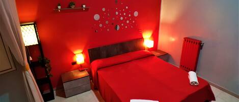 A room with a red bedspread, two bedside lamps, a wall-mounted shelf with plants, a radiator, and a tiled floor.