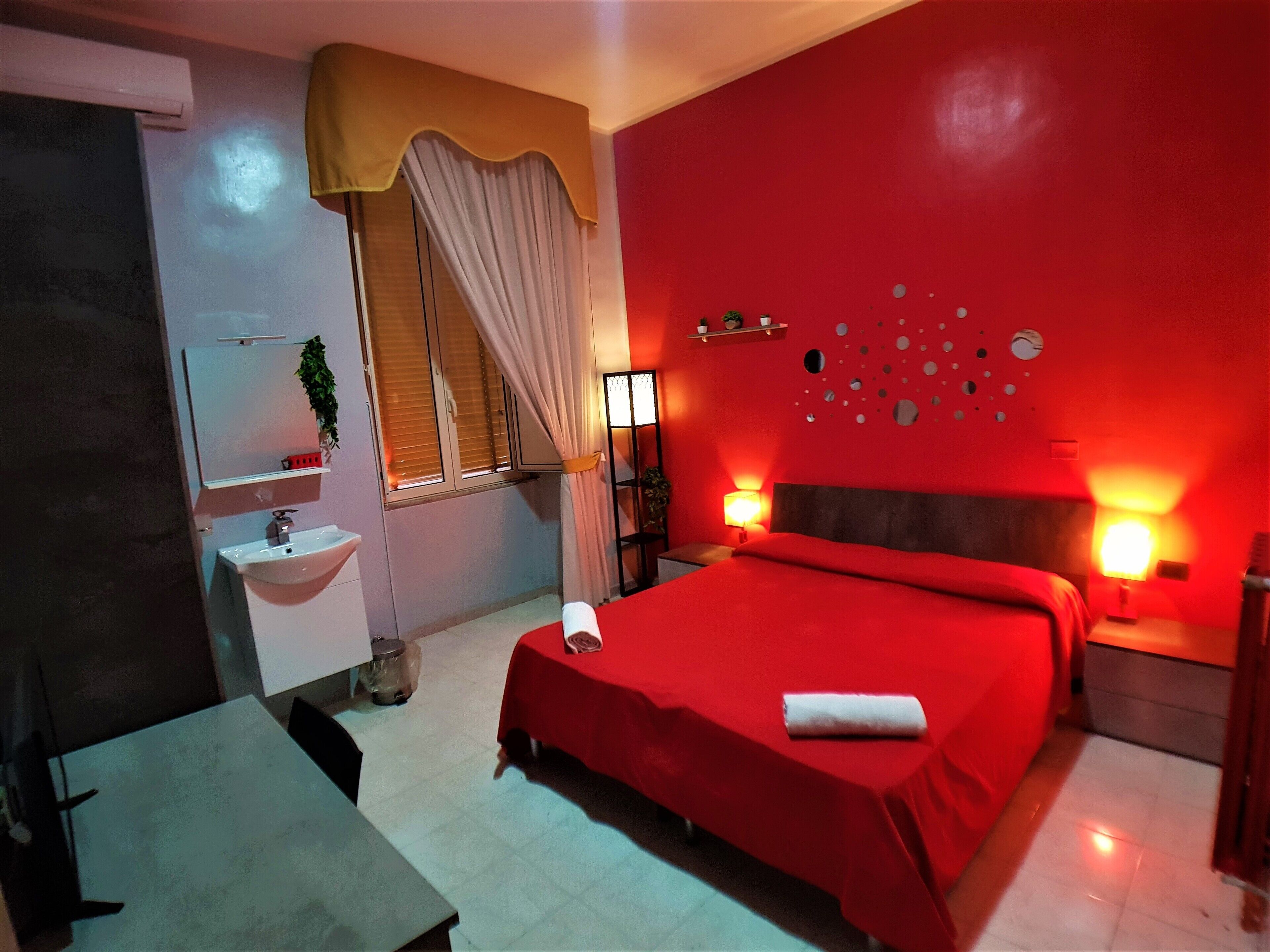 A hotel room with a red bedspread, a bedside table with a lamp, a large mirror, a window with curtains, and a wall with circular decorations.