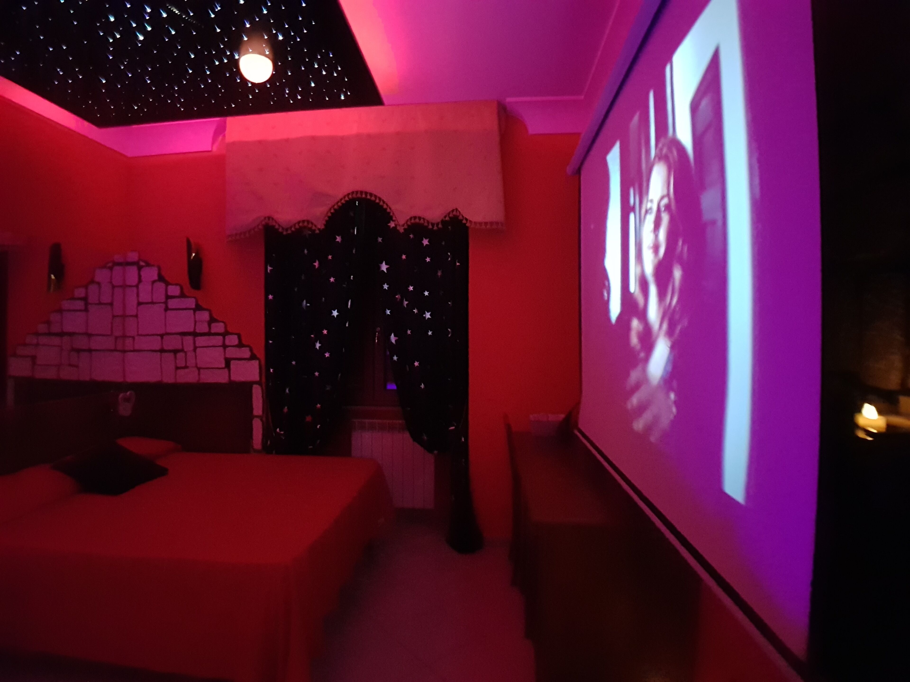 Ein Zimmer mit Projektionsleinwand, auf der ein Film läuft, einem Bett, einem kleinen Tisch und einer Wand mit Backsteinmuster.