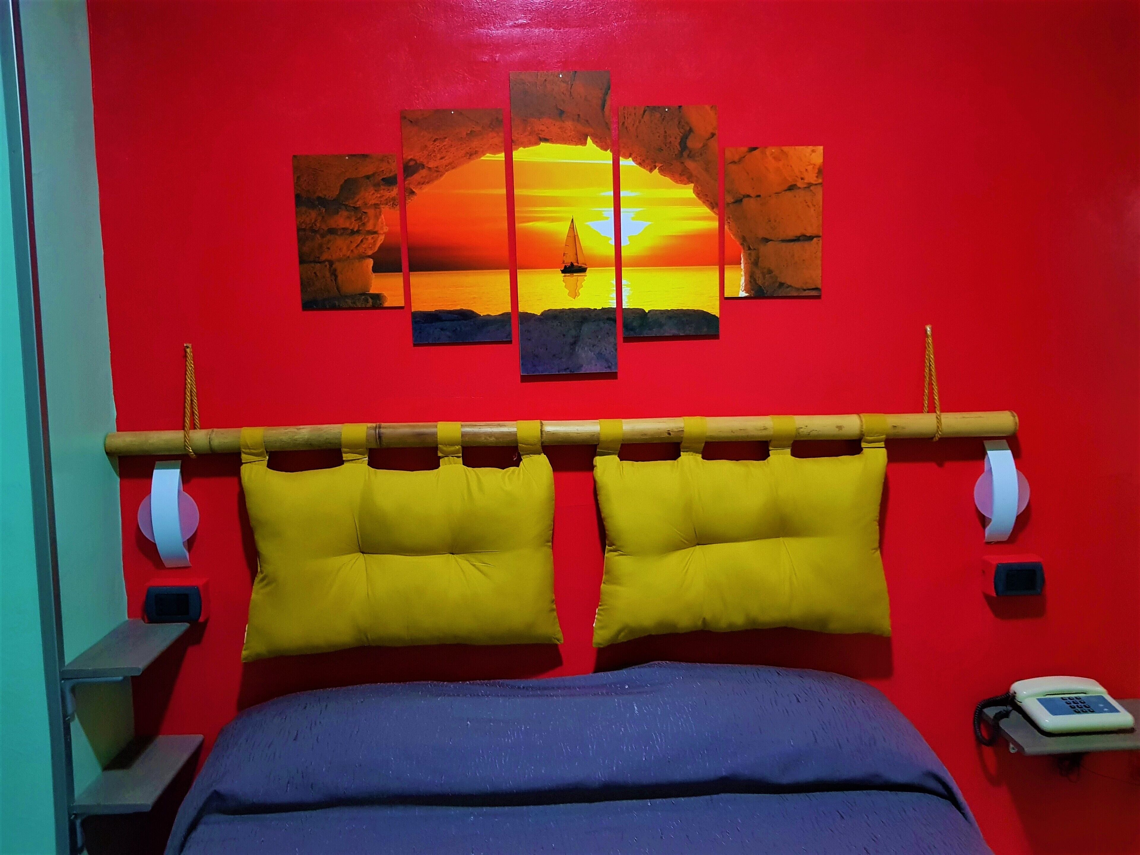 A room with a red wall, a bed with yellow pillows, a wooden headboard, and a painting of a sunset.