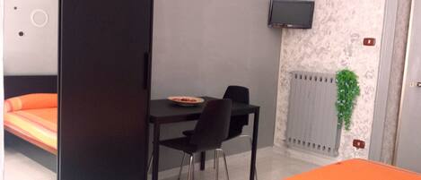 A room with a bed, a table with chairs, a TV mounted on the wall, and a radiator.