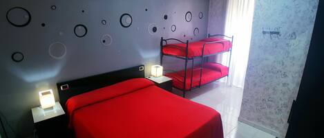 Una habitación de hotel con cama roja, literas, una mesita de noche con lámpara y una pared con diseños circulares.