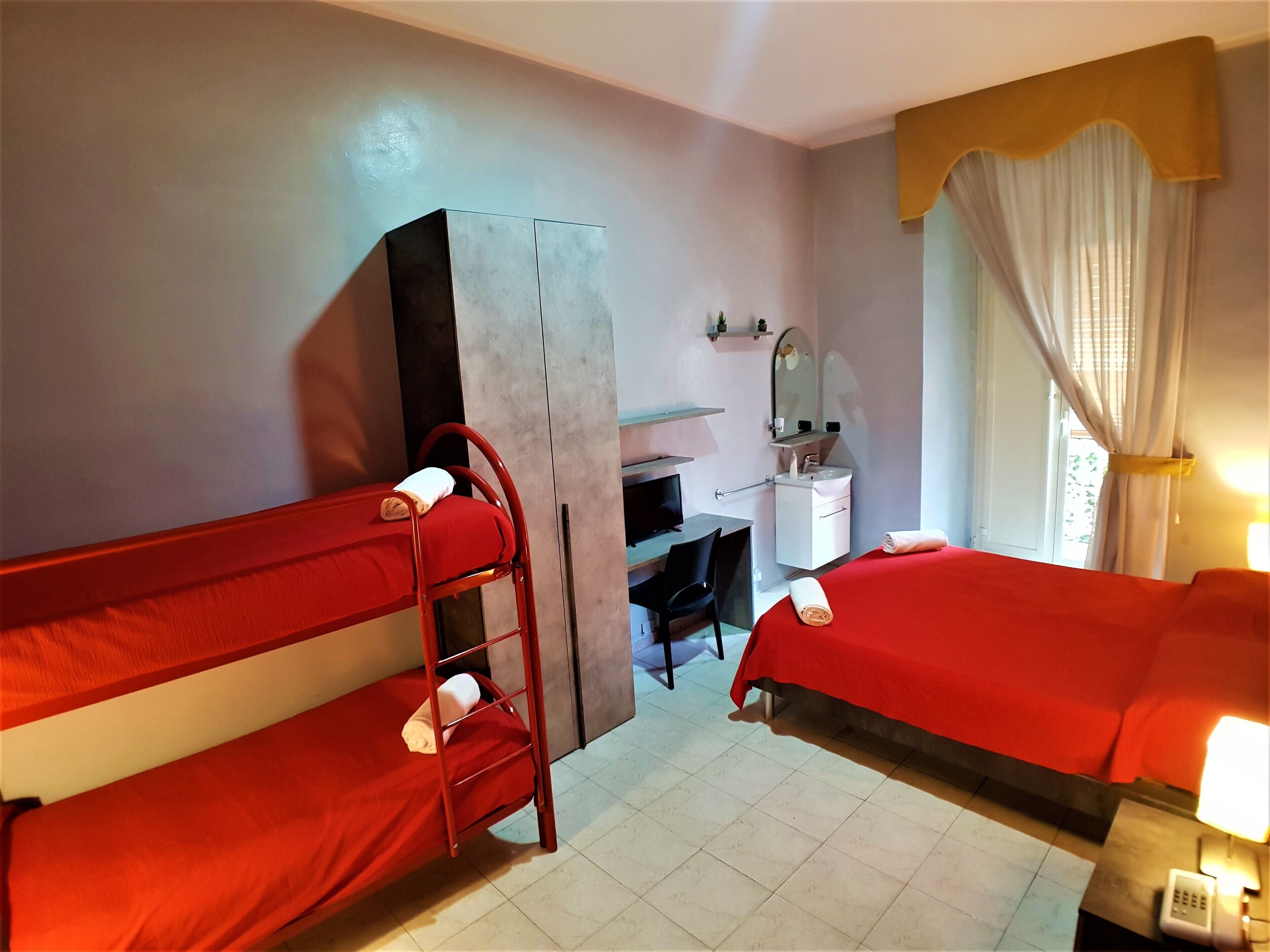 A hotel room with two bunk beds, a desk with a chair, a small cabinet, a mirror, and a window with curtains.