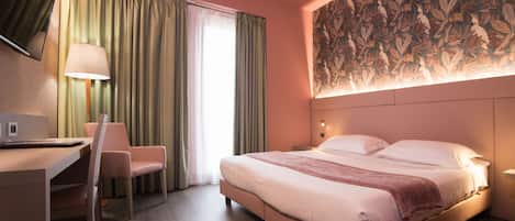 A hotel room with a bed, a desk with a chair, a TV, and a large floral-patterned wall.