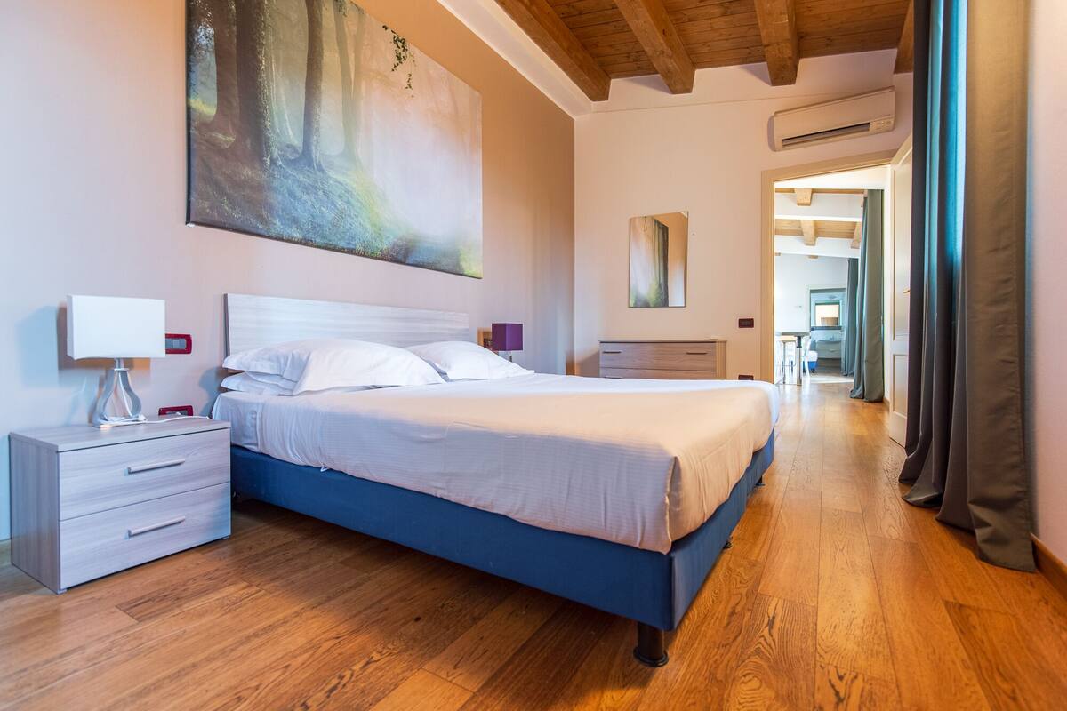 A bedroom with a large bed, a nightstand, a dresser, and a painting on the wall.