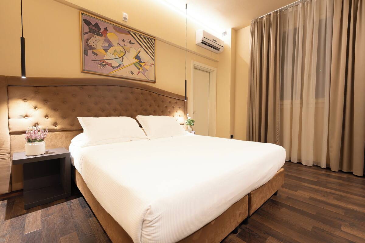 A neatly made bed in a hotel room with a tufted headboard, a bedside table with a vase of flowers, and a large abstract painting on the wall.